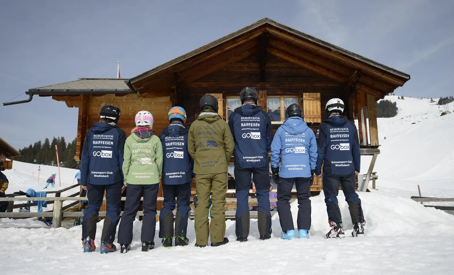 Clubmitglieder in Vereinsjacken von hinten vor einer Berghütte